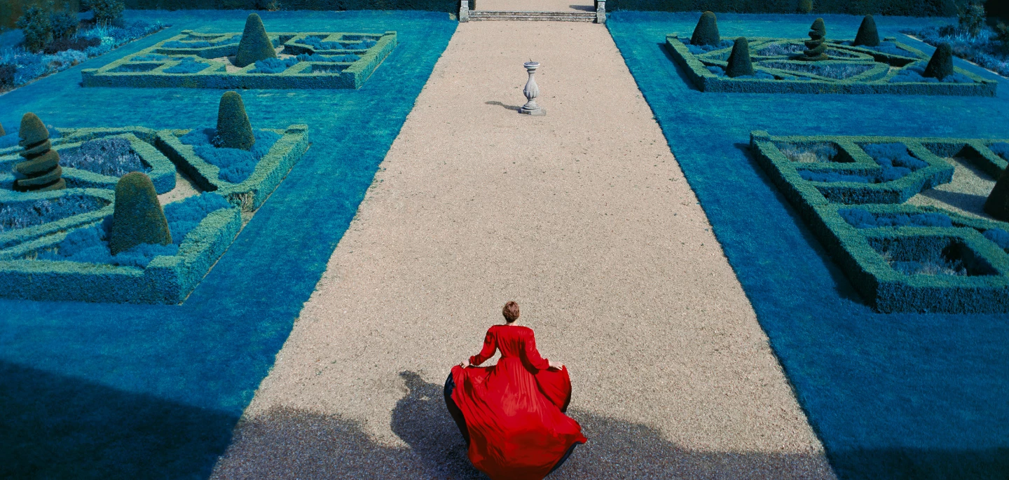 Aerial view of a formal garden with intricate geometric hedges and pathways. In the center, a person wearing a flowing red dress stands with their back to the viewer, facing a stone statue in the distance. The garden is surrounded by vibrant blue-green foliage, creating a striking contrast with the red dress.