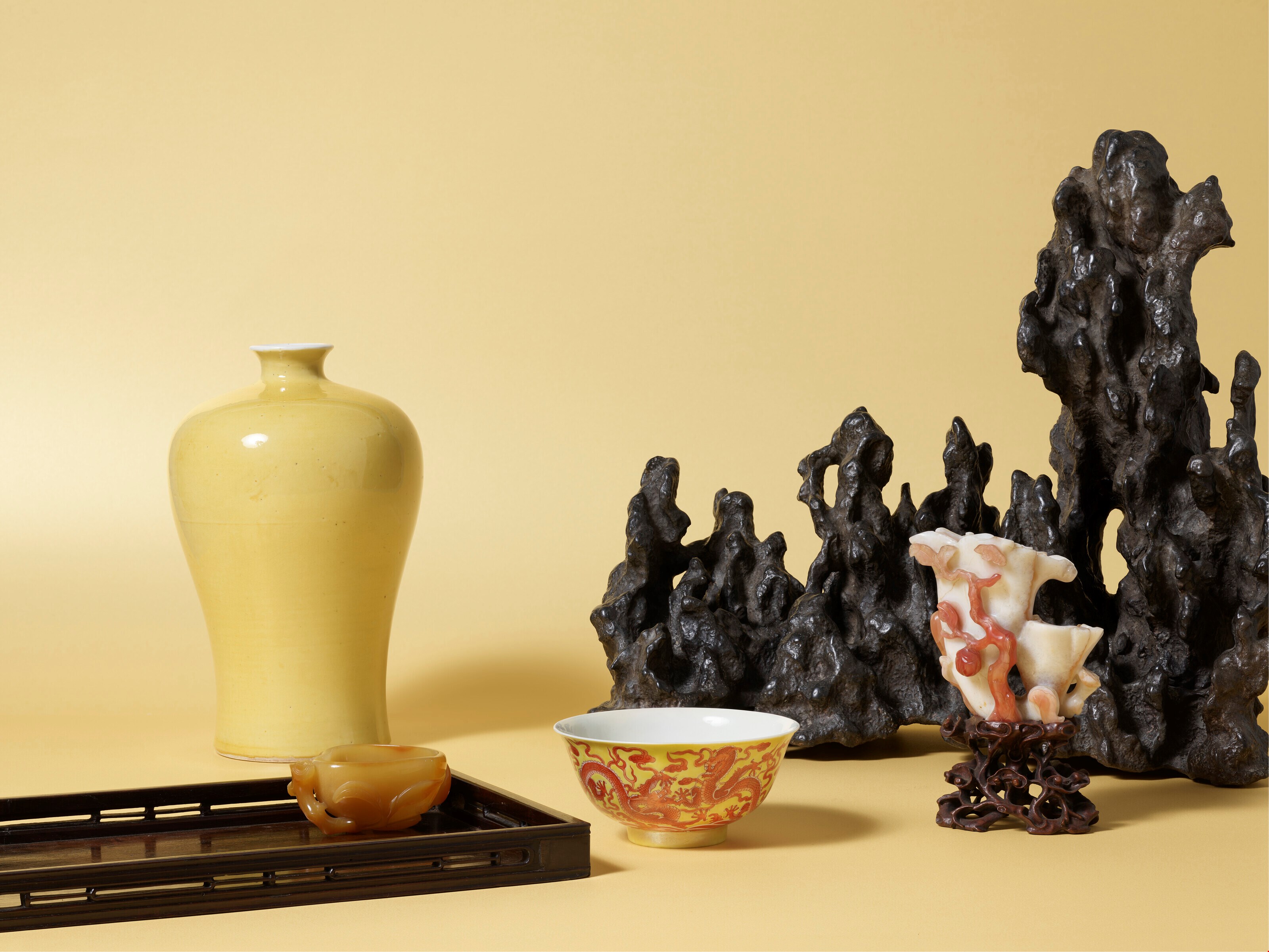 A collection of exquisite decorative objects arranged on a soft yellow background. The display features a tall, elegant yellow vase, a delicately painted ceramic bowl, and a small carved piece, all complemented by a striking, textured rock formation that adds depth and contrast to the composition.